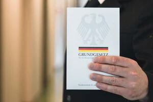Auf dem Bild wird das Grundgesetz in Form eines Buches gezeigt, das eine Person vor dem Oberkörper in der Hand hält. Die Person ist nicht vollständig zu sehen.